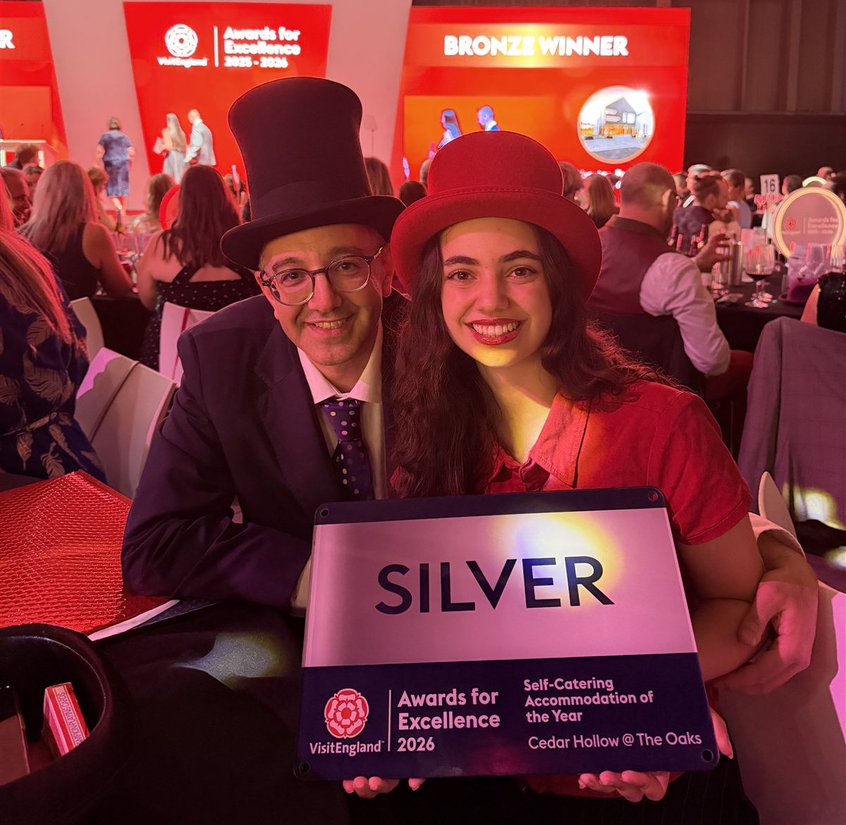 Dr Yaz in a black top hat celebrating at the VisitEngland Awards for Excellence 2026, holding the Silver award for Self-Catering Accommodation of the Year won by Cedar Hollow @ The Oaks