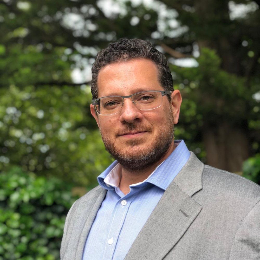 Roi Amir smiling in a grey blazer and blue shirt, photographed outdoors among trees