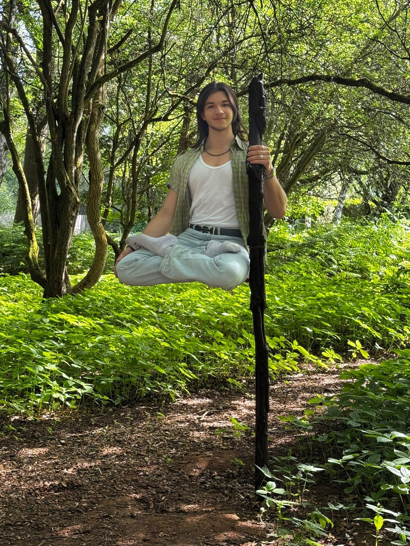 A young man sits cross-legged in mid-air in a sunlit woodland, holding a tall wooden staff, with no visible support beneath him