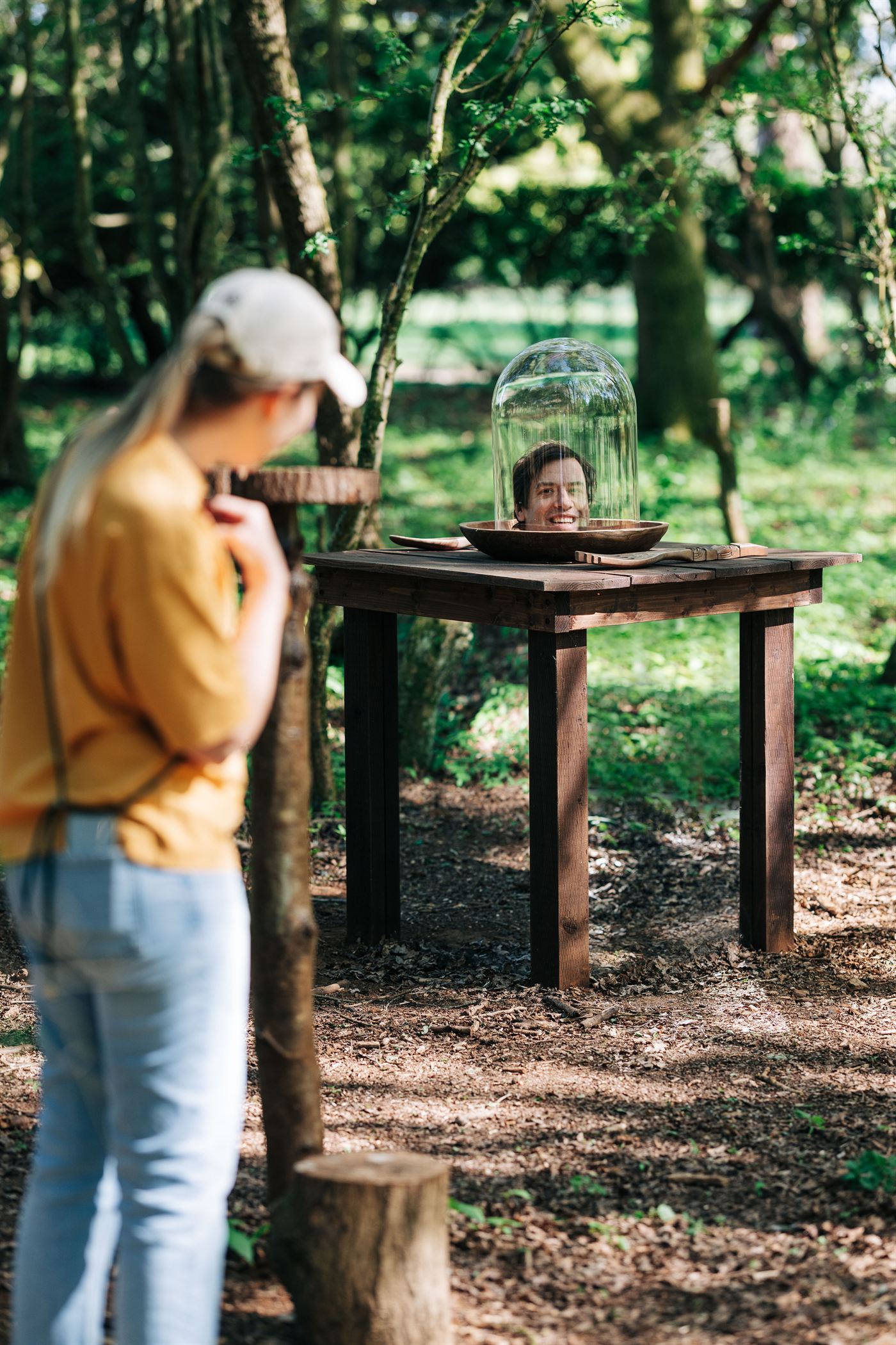 A disembodied head appears under a glass dome on a wooden table in the woods