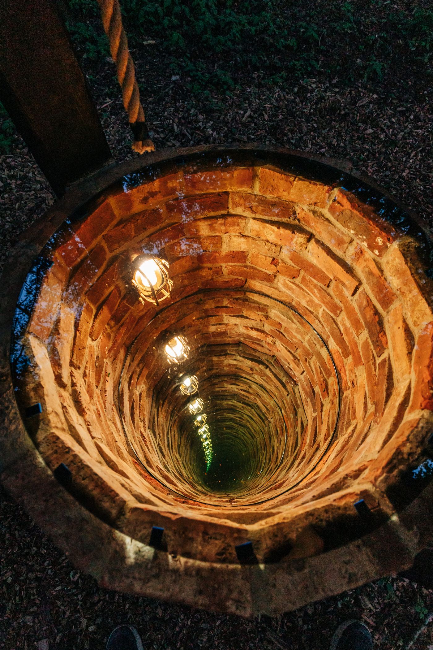 Looking down into a deep brick well that appears to descend forever, lit by glowing lanterns